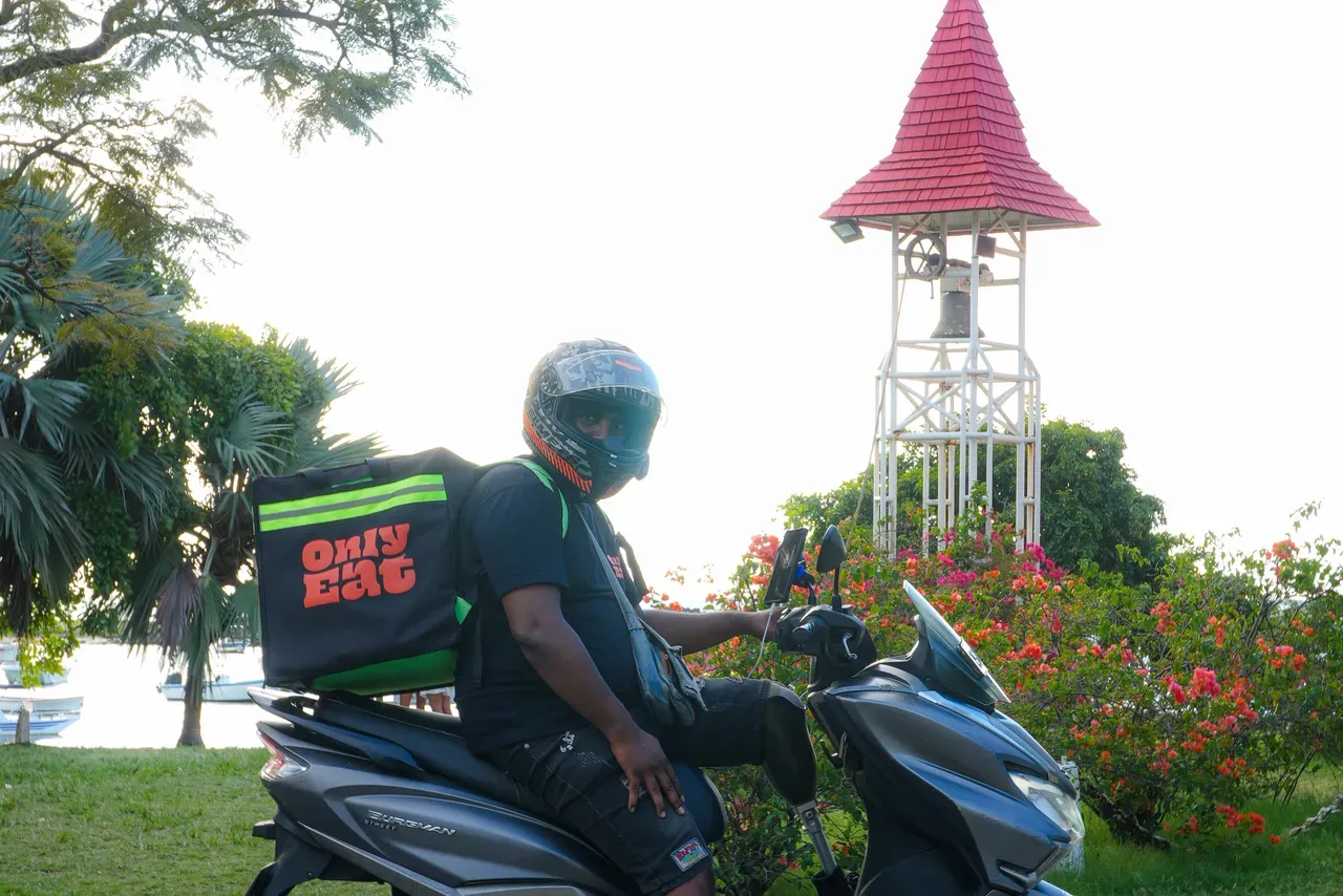An OnlyEat rider on a scooter delivering past the red-roofed church of Cap Malheureux, Mauritius.